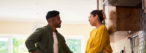 A man and woman are standing in a kitchen, with the woman sitting on the counter and the man standing next to her.