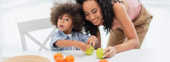 A woman and a child are sitting at a table with oranges and apples.
