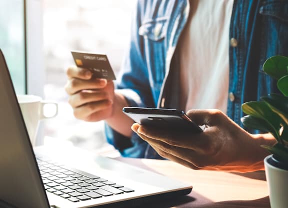 A person is holding a credit card and a smartphone while sitting in front of a laptop.