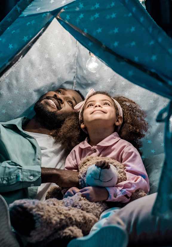 A man and a little girl are lying in a tent with a teddy bear.