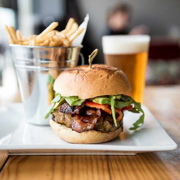 A burger with bacon and lettuce on a bun with a glass of beer in the background.