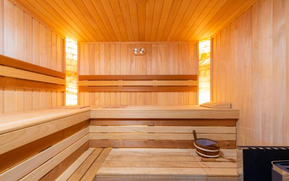 A wooden sauna with a glass door is surrounded by plants and has a towel on the side.
