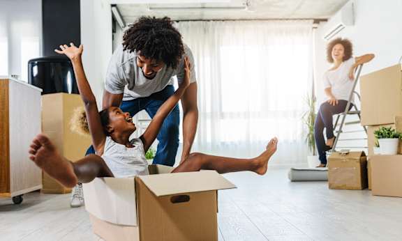 A man and a child are playing with a cardboard box in a room with other boxes and a woman sitting on a chair in the background.