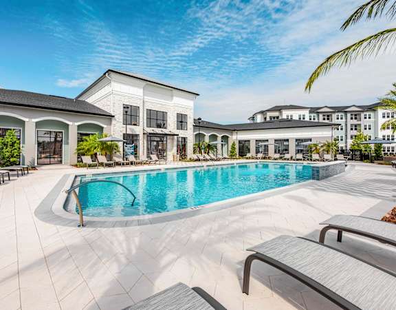 A large swimming pool is surrounded by lounge chairs in front of a white building.