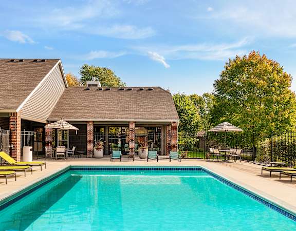 A swimming pool in front of a house with a patio and lawn chairs.