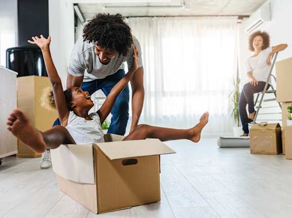 A man and a child are playing with a cardboard box in a room with other boxes and a woman sitting on a chair in the background.