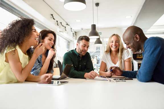 A group of people are gathered around a table, looking at a laptop screen and discussing something.