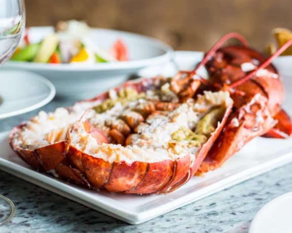 A lobster is served on a white plate with a glass of wine and a bowl of salad in the background.