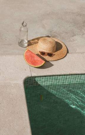 A straw hat and a slice of watermelon are placed on the edge of a pool.