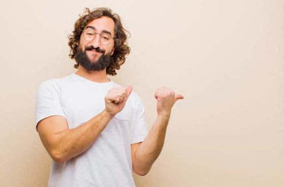 A man with curly hair and a beard is wearing glasses and a white t-shirt, and he is making a gesture with both hands.