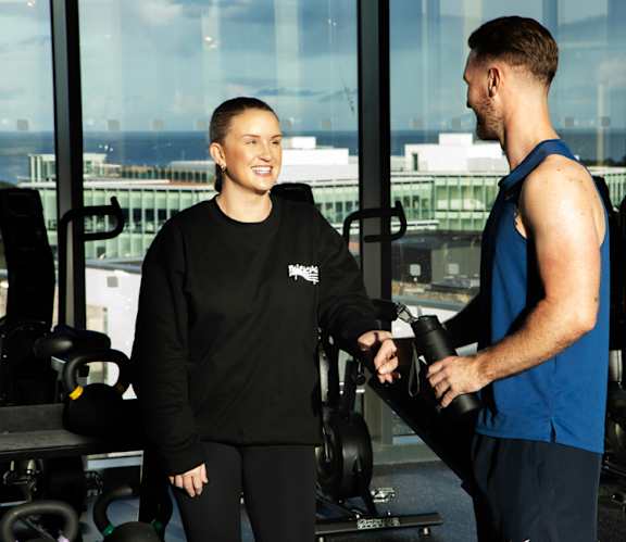 A man and a woman in a gym, the woman is wearing a black hoodie with a logo on it.