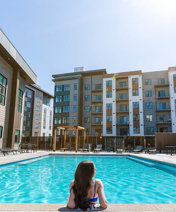 A woman sits by a pool in front of a large apartment building.