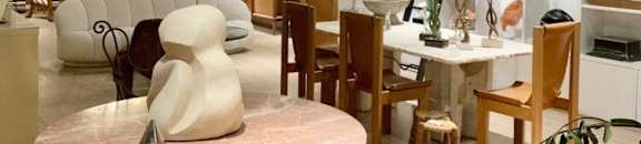 A round table with a green base and a pink marble top is in the center of a room with a white wall and a shelf with various items on it.