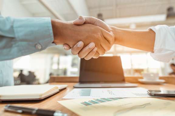 Two people shaking hands over a desk with documents and a laptop.