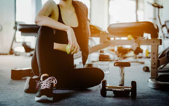 A woman is sitting on the floor in a gym, holding a water bottle and looking at her phone.