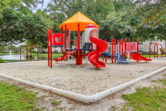 Blue Red and Yellow Playground on Sand with Building Exterior and Treeline in the Background