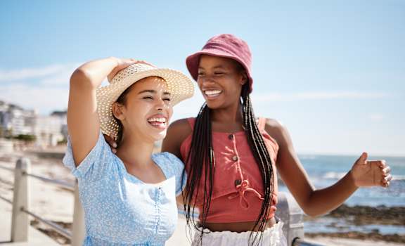 Two women wearing summer clothes are standing by the sea.