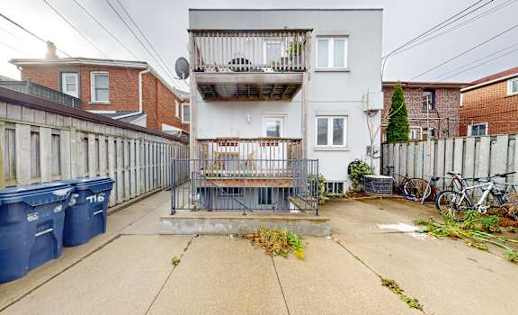 A grey building with a balcony is in the middle of a courtyard with blue bins and a fence.