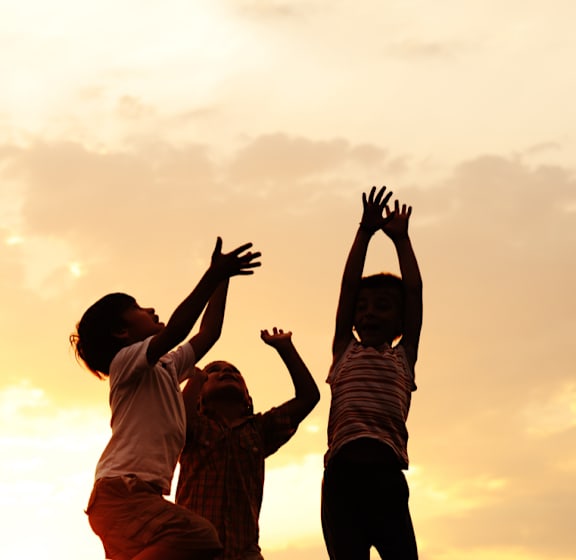 Three children are silhouetted against a sunset sky as they playfully reach for the sky.