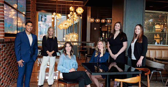 A group of people are sitting and standing in a room with a brick wall and a glass table.
