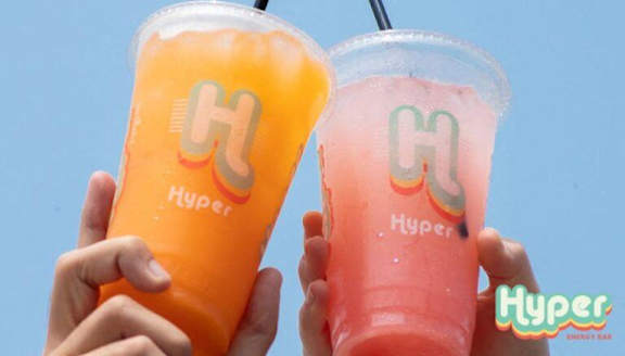 Two cups of Hyper drinks are being held up against a blue sky.