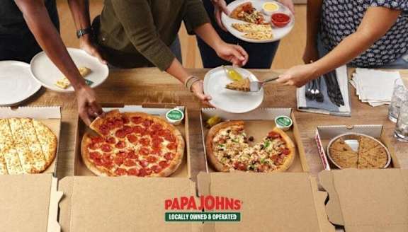 A group of people are gathered around a table with various pizzas from Papa John's.