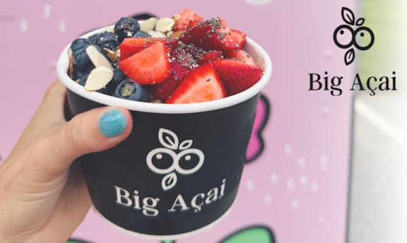 A hand holding a cup of Big Acai with a strawberry and blueberry smoothie topped with nuts.