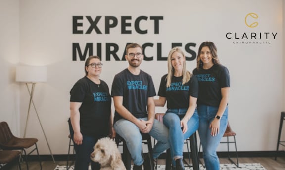 Four people wearing black shirts with "Expect Miracles" printed on them are standing in front of a wall with the same text.