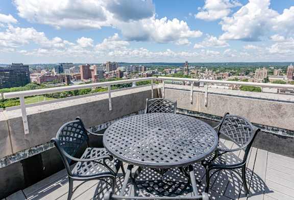 A table and chairs are set up on a balcony with a cityscape in the background.