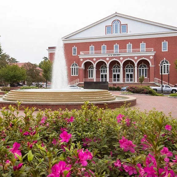 A fountain in front of a red building with a woman walking in the background.