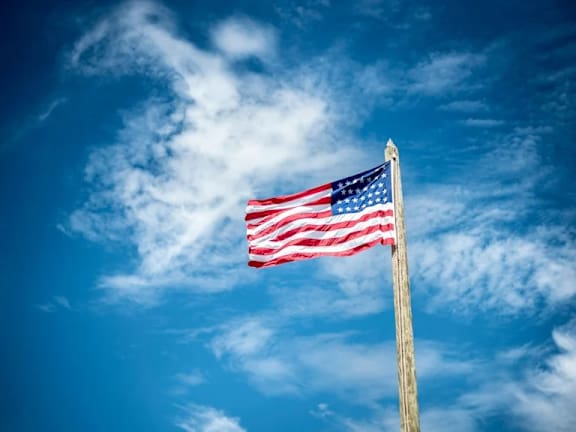 The American flag is flying high on a wooden pole against a blue sky with clouds.