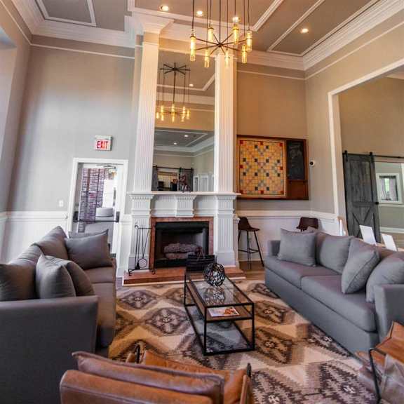 The amazing resident lounge with gray couches, decorations, large windows, unique overhead lighting, a fireplace, brown leather chairs, and coffee tables at Sycamore Farms Apartment Homes. 