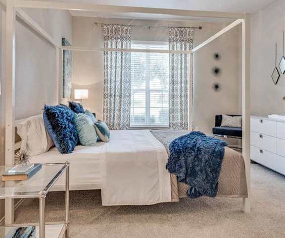 A spacious bedroom with a queen-size bed with a white and blue bedspread, a dresser, wall art, lamps, decorations, overhead lighting with a fan, window, and nightstands at Sycamore Farms Apartment Homes. 