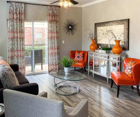 The spacious living room with a couch, throw pillows, large windows, wall art, red and gray plush chairs, a glass coffee table, decorations, a TV, and glass doors to the balcony at Sycamore Farms Apartment Homes. 