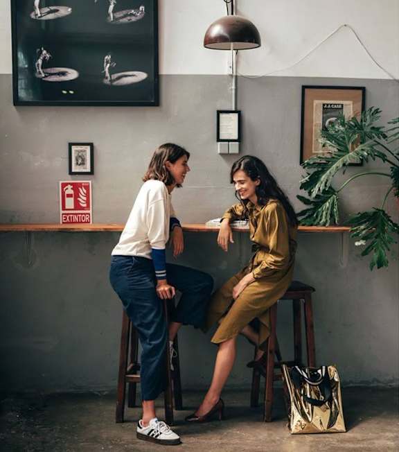 Two women sitting at a table in a room with a plant and a poster on the wall.
