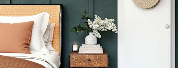 A bed with a white pillow and a brown pillow case sits next to a nightstand with a plant on it.