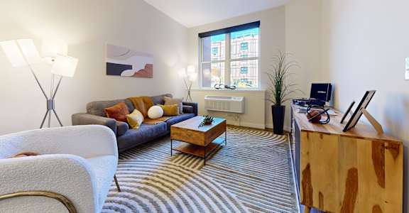A living room with a grey couch, a wooden coffee table, a striped carpet, and a window with a view of a building outside.