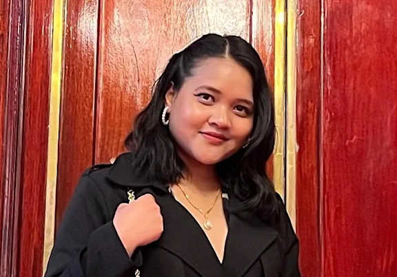 A woman with a black blazer and a gold necklace is standing in front of a red wall.