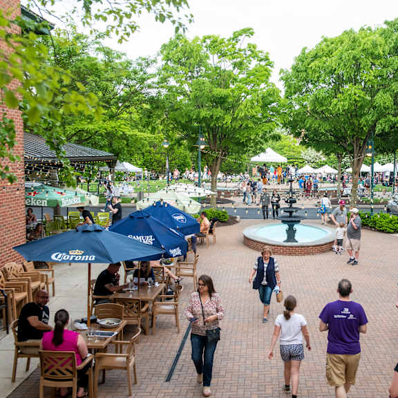 A group of people are walking around a courtyard with tables and umbrellas.