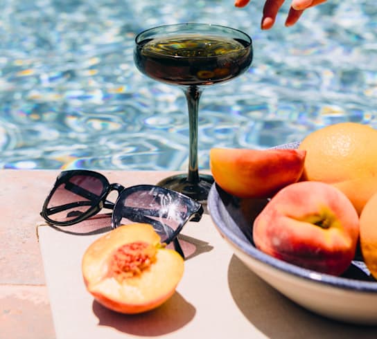 A martini glass, a bowl of fruit, and sunglasses are on a table by a pool.