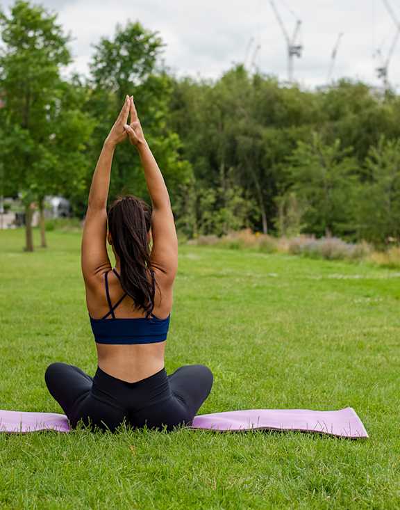 A woman in a blue sports bra and black leggings is sitting on a pink yoga mat in a park, performing a yoga pose.