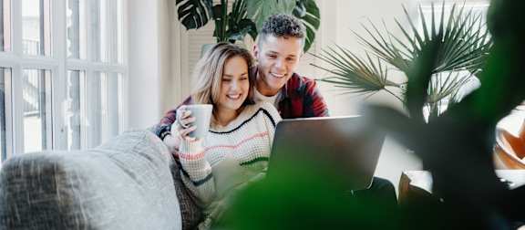 A man and a woman are sitting on a couch with a laptop and a cup in their hands.