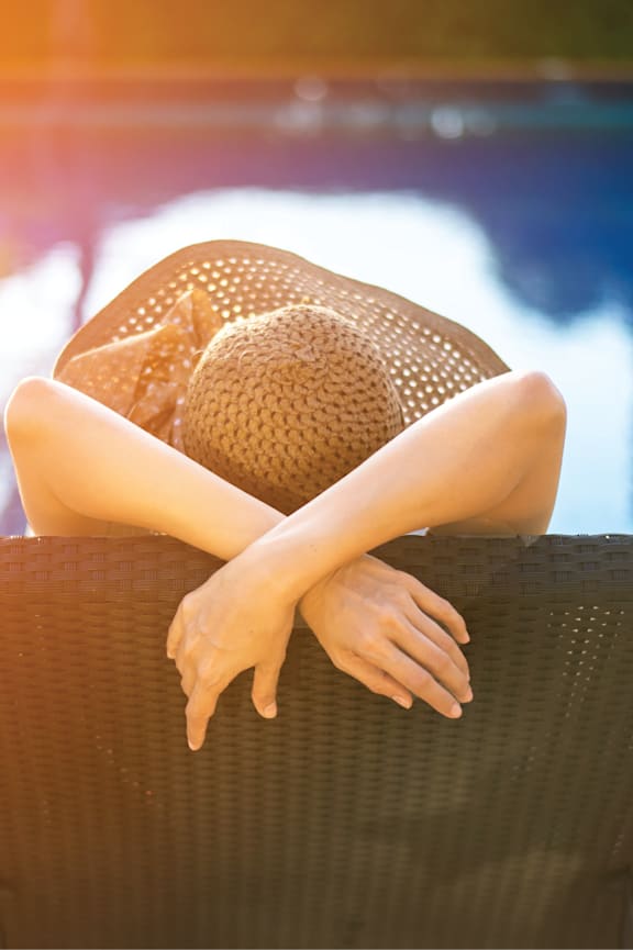 A woman in a straw hat is sitting on a chair with her arms resting on the back of the chair.