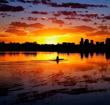 A person is kayaking on a calm body of water with a city skyline silhouette in the background during a sunset.