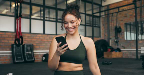 A woman in a gym is sitting on the floor and looking at her phone.
