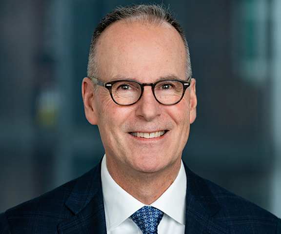 A man with glasses and a blue tie is smiling.