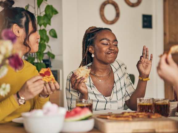 Two women are eating pizza at a table with a watermelon and a vase of flowers.