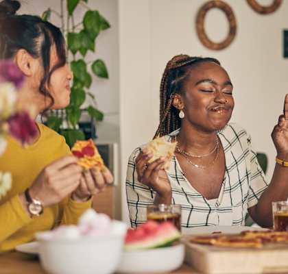 Two women are eating pizza at a table with a watermelon and a vase of flowers.