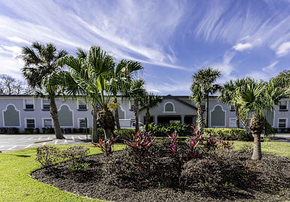 A white building with arched windows is surrounded by a well-manicured garden with palm trees.
