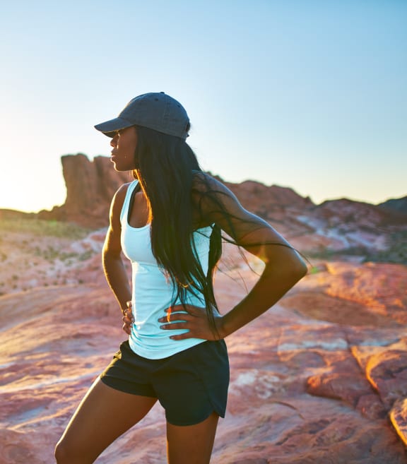 a woman in a white tank top and black shorts stands on a rock as the sun sets
